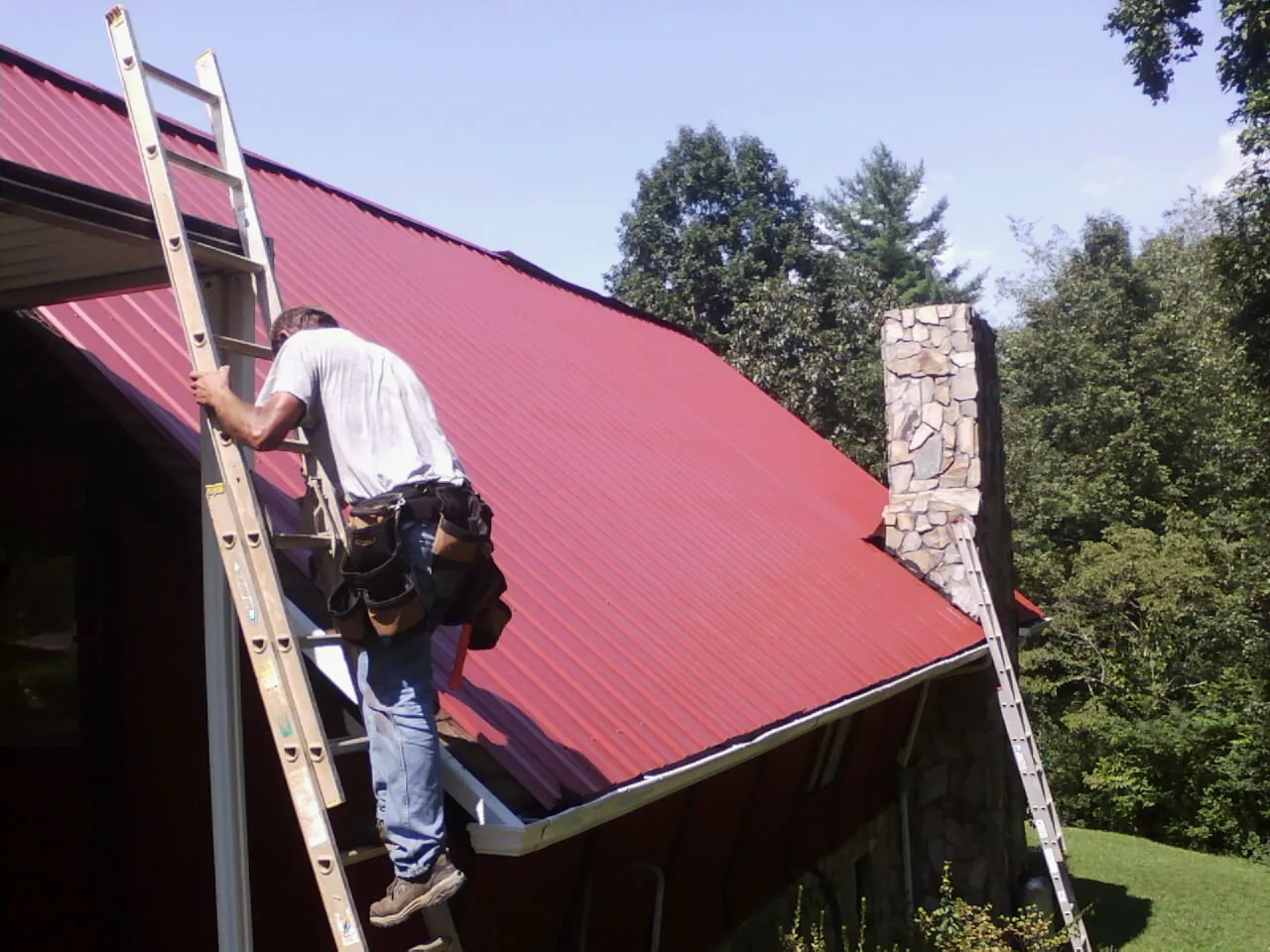 Roofer on a ladder beside a red metal roof and stone chimney — Soffit & Fascia Repair by Roofing Brothers in Golf